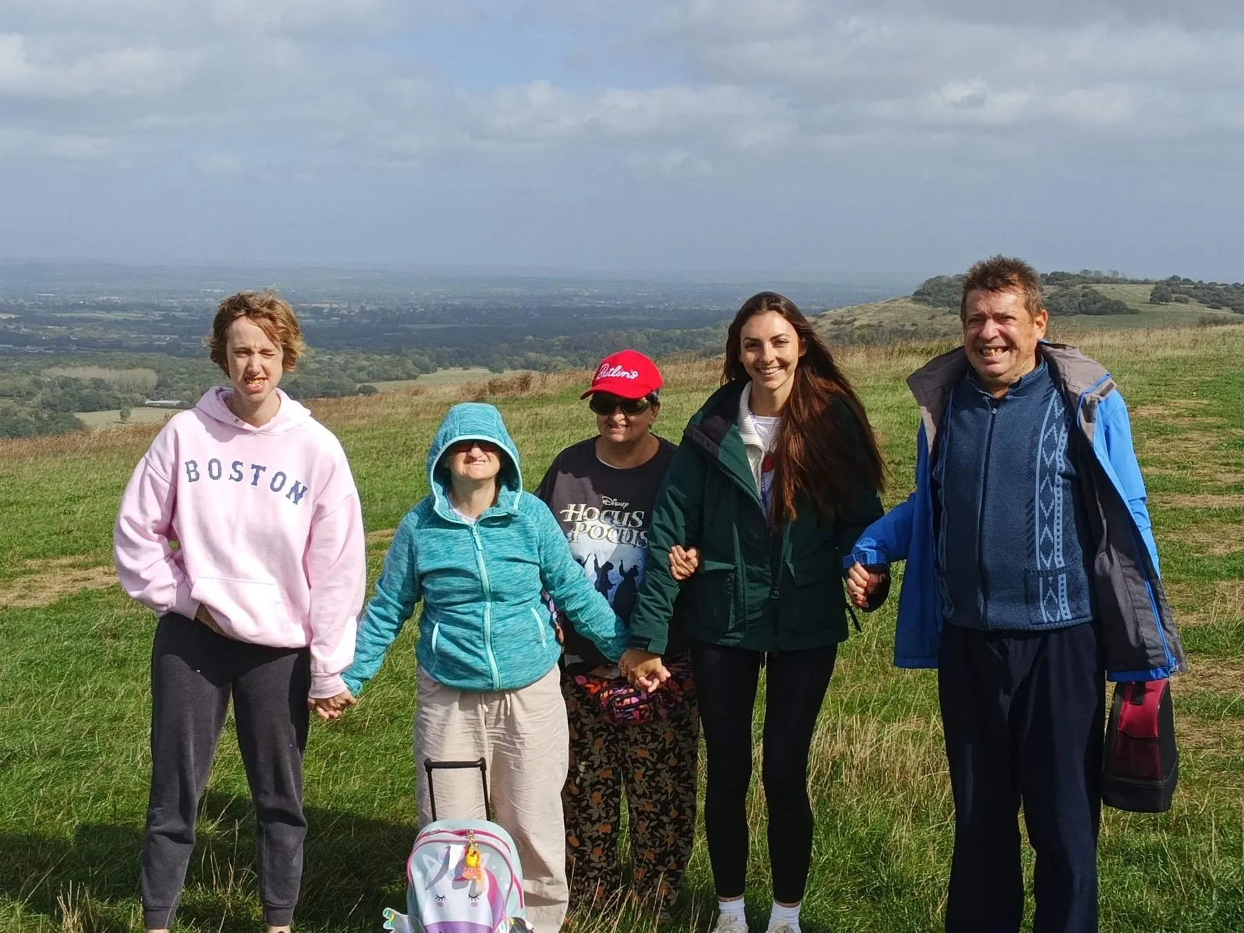 Five people stand in a row holding hands on top of the Downs, smiling, with fields stretching out below