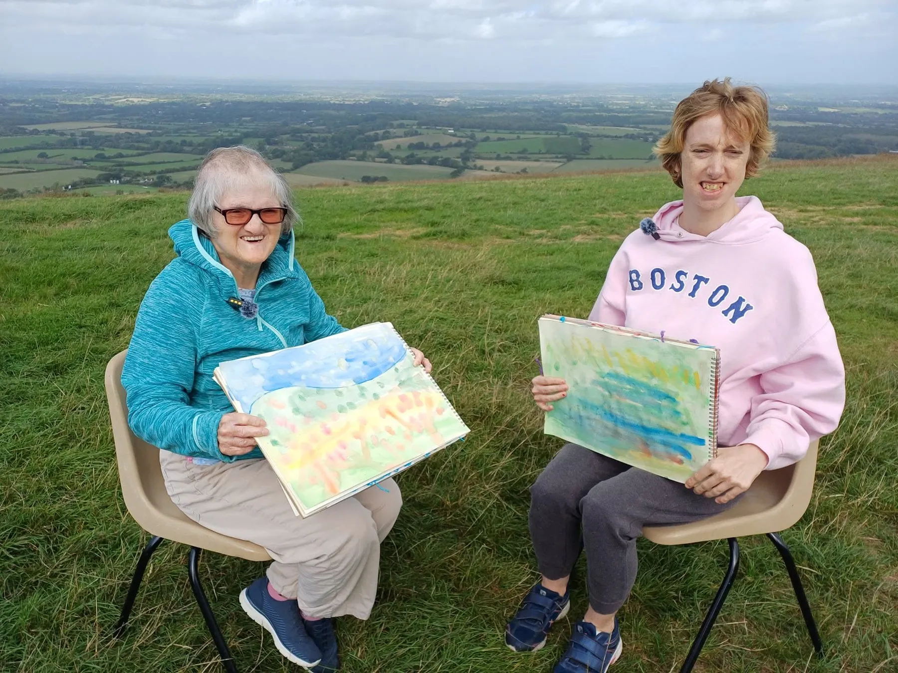 Two women sit on chairs on the grass at the top of the Downs, each holding up a watercolour painting of the view