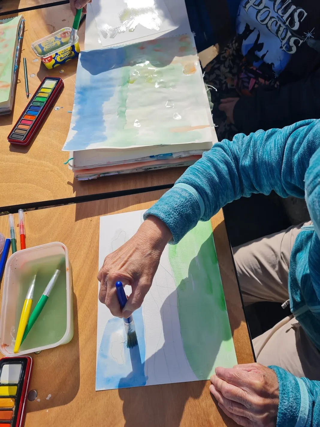 Close-up of a hand painting blue sky in watercolour, with paint boxes and a water pot on the table